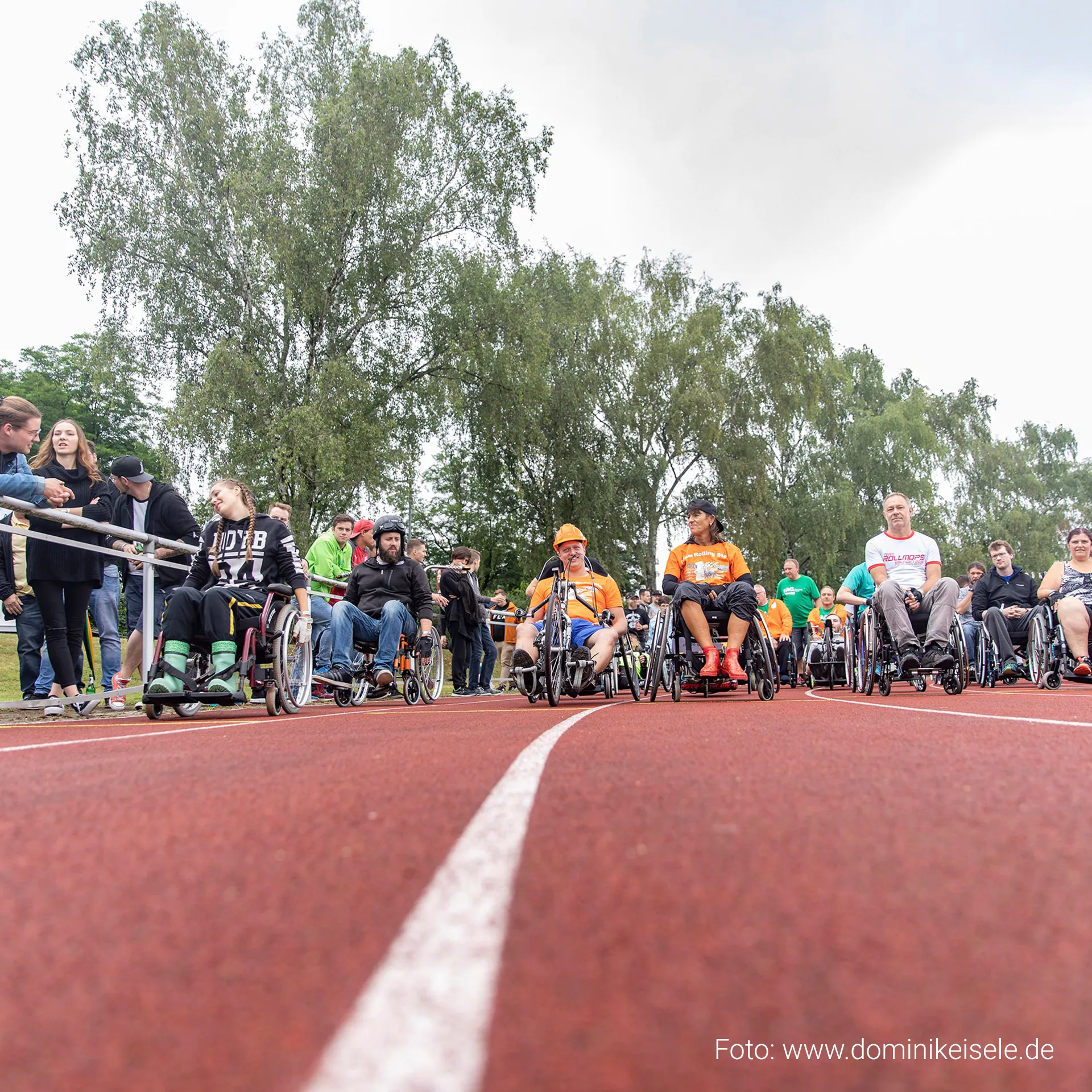 Teilnehmende in Rollstühlen auf der Laufbahn eines inklusiven Sportevents im Eichenbachstadion Eislingen. Zuschauerinnen und Zuschauer verfolgen das 24-Stunden-Rollstuhlrennen, das Begegnung, Bewegung und gesellschaftliche Teilhabe fördert. Bild von Fotograf Dominik Eisele.